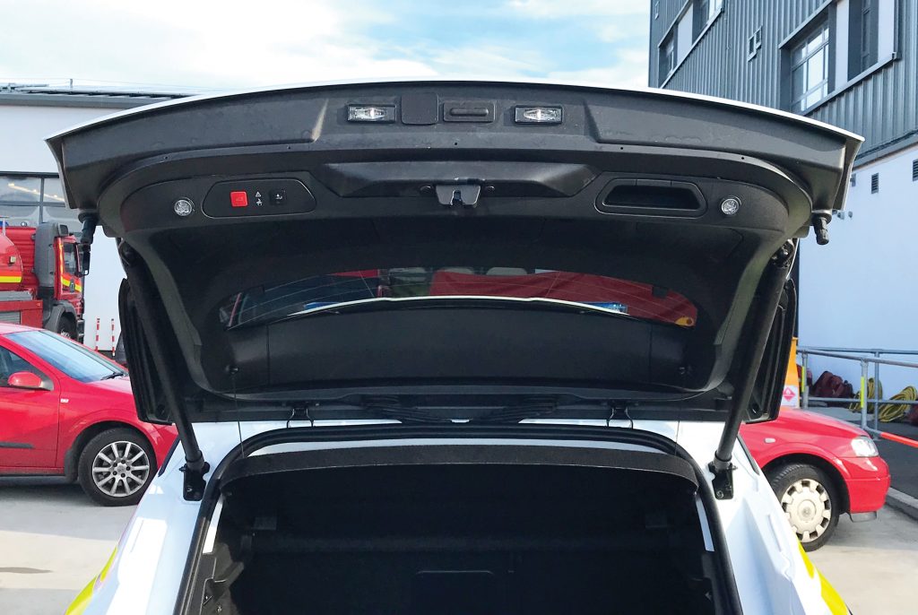 Back view of white fire response vehicle with open boot on cemented vehicle forecourt in front of various vehicles and shutter doors