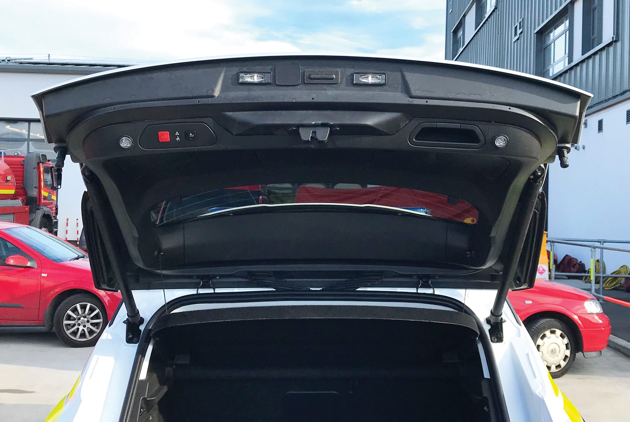 Back view of white fire response vehicle with open boot on cemented vehicle forecourt in front of various vehicles and shutter doors