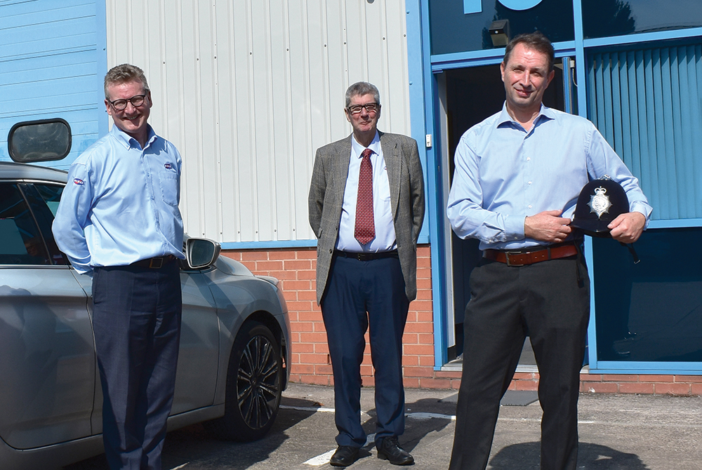 Three smiling corporate men stand in front of building unit with car. One man holds police helmet